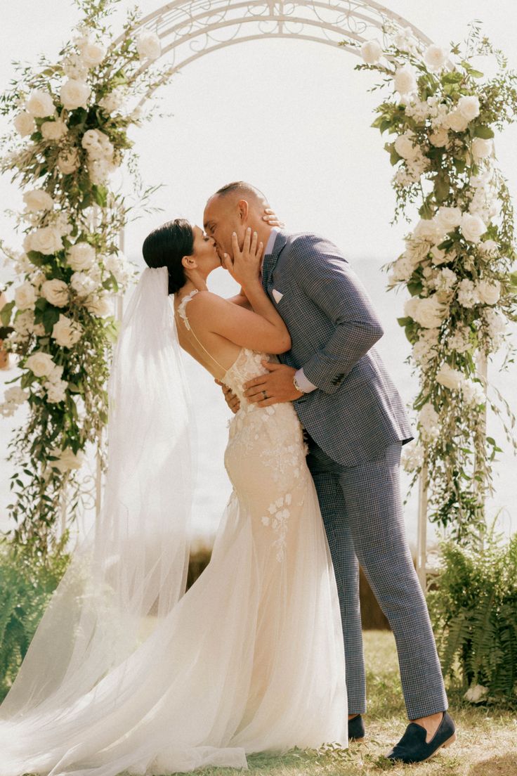 a bride and groom kissing under an arch decorated with white flowers at their wedding ceremony