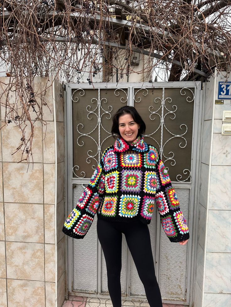 a woman standing in front of a door wearing a colorful crocheted jacket