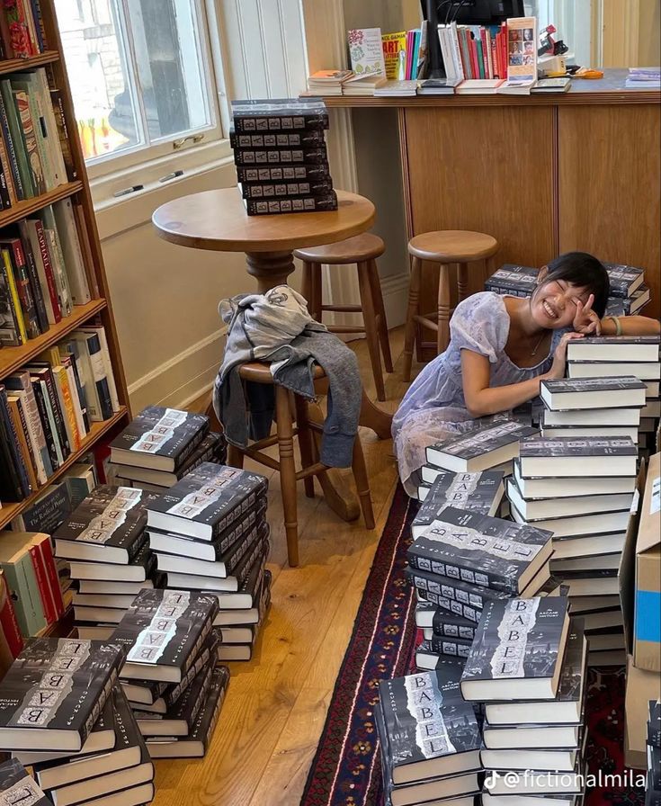 a woman is laying on the floor surrounded by stacks of books in a room full of tables and chairs