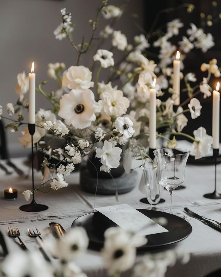 the table is set with white flowers and black place settings, silverware and candles