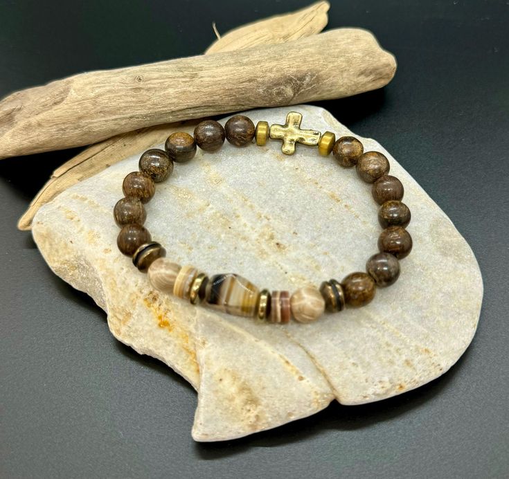 Introducing the Earthbound Valor bracelet, handcrafted by JP Designs. This rugged bracelet combines the grounding properties of bronzite with the organic textures of shell heishi beads, the protective energy of pyrite, and the deep hues of smoky quartz. The addition of bronze accents and a central cross charm symbolizes strength, faith, and resilience, making this piece not only stylish but also meaningful. Perfect for the modern man who values quality craftsmanship and the power of natural elem Spiritual Brown Beaded Bracelets With Gemstone Beads, Spiritual Brown Gemstone Beaded Bracelets, Artisan Brown Bracelet With Gemstone Beads, Artisan Brown Bracelets With Gemstone Beads, Spiritual Brown Gemstone Beads Bracelets, Meditation Brown Bracelet With Polished Beads, Earthy Brown Bracelets With Gemstone Beads, Earthy Brown Gemstone Beads Bracelets, Handmade Brown Stretch Bracelet For Meditation