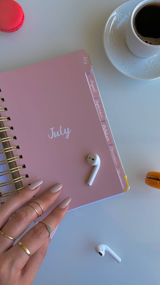 a woman's hand holding an open pink notebook next to ear buds and coffee