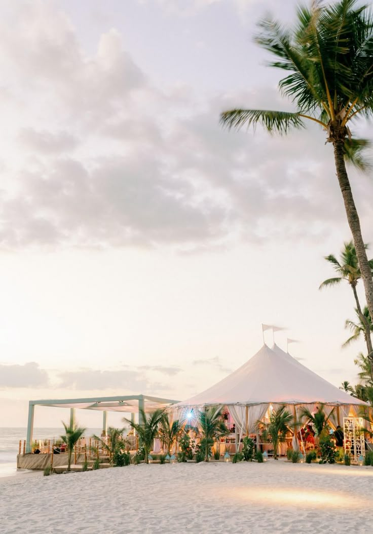 a tent set up on the beach with palm trees in front of it at sunset