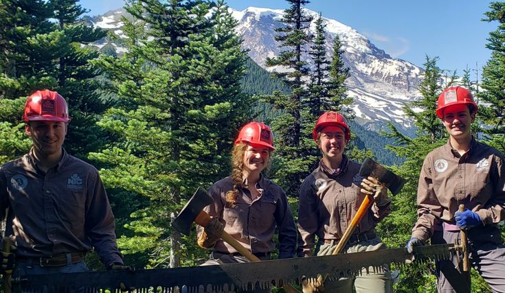 four people wearing hard hats and holding shovels standing in front of a fence with mountains in the background