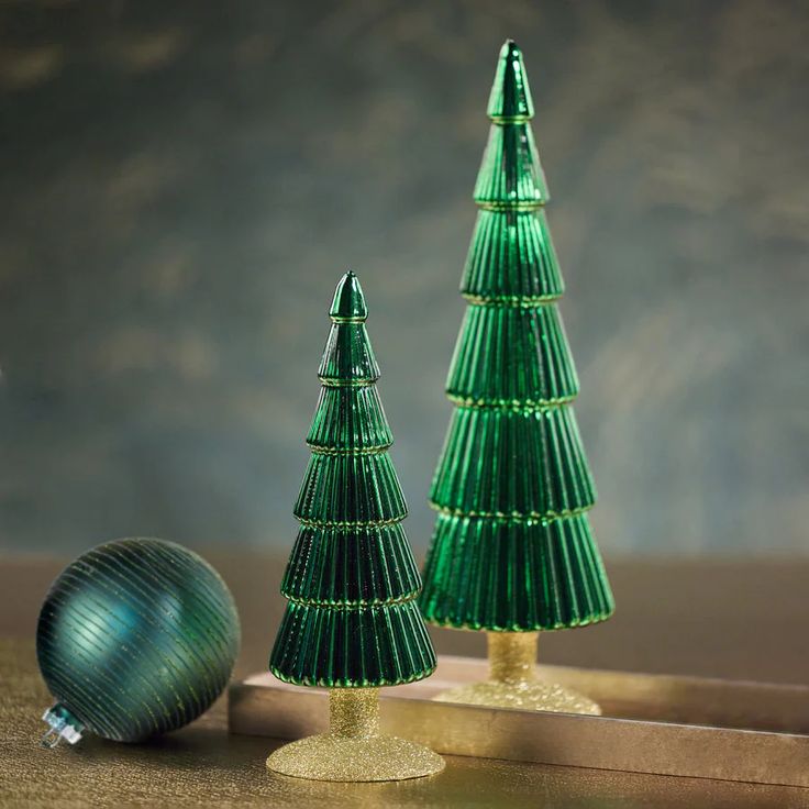 two green glass christmas trees sitting on top of a wooden table next to a silver ornament