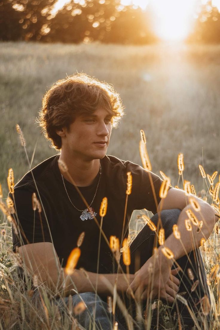 a young man is sitting in the grass with his hands on his knees and looking off into the distance