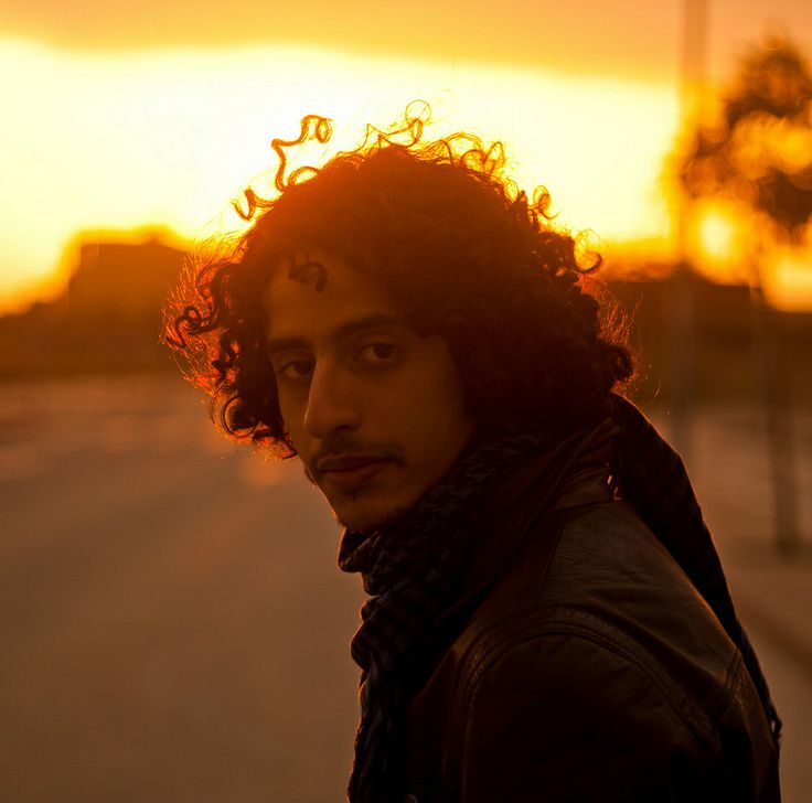 a young man with curly hair standing on the street at sunset, looking into the camera