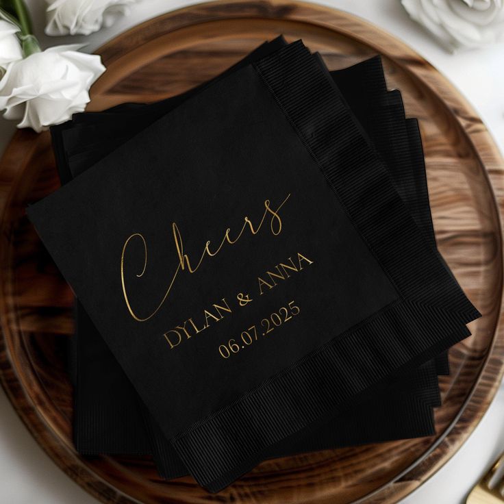 black napkins with gold foil lettering on them sitting on a wooden plate next to white flowers