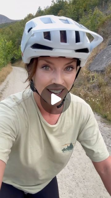 a woman riding a bike down a dirt road next to a lush green forest covered hillside