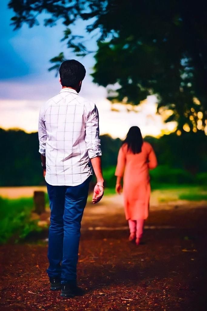 a man and woman walking down a dirt road at dusk with trees in the background