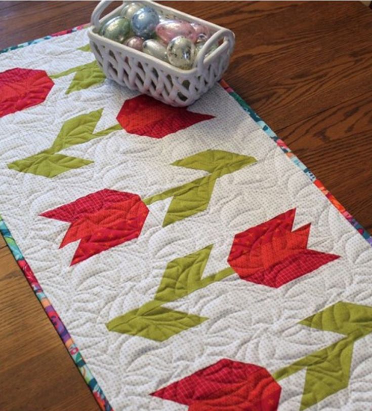 a quilted table runner with red and green leaves on it, next to a basket filled with eggs