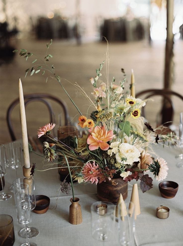 an arrangement of flowers on a table with candles and wine glasses in the foreground