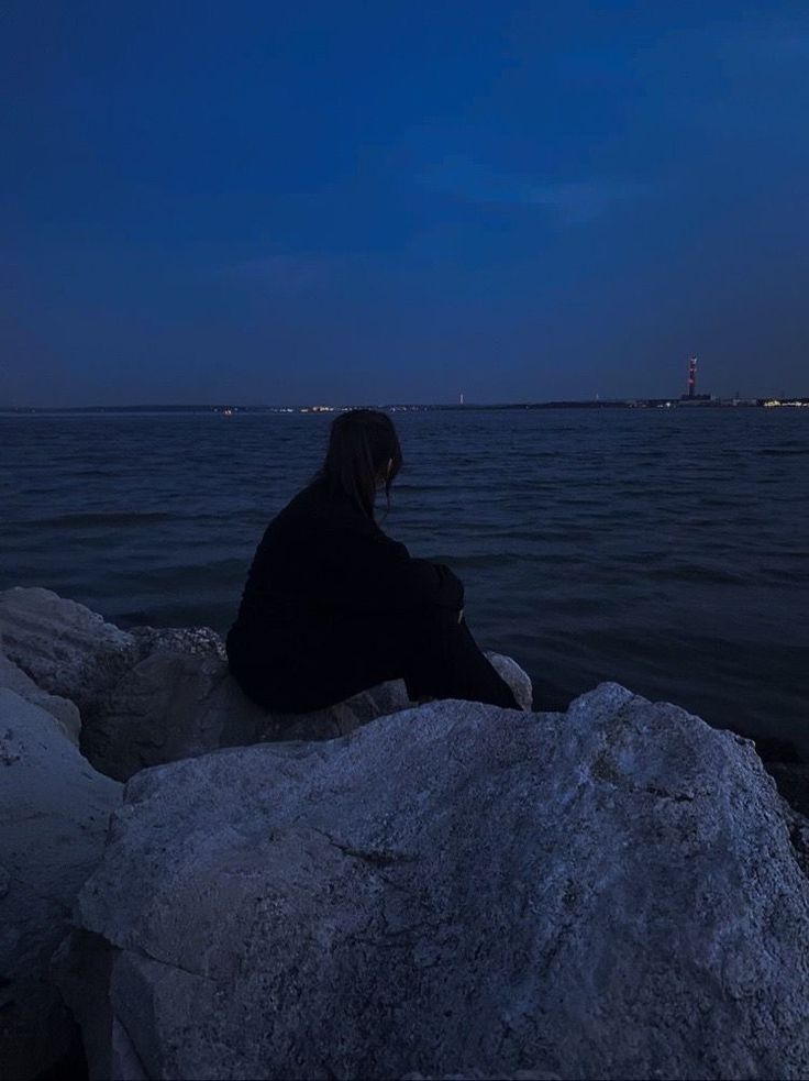 a person sitting on top of a rock next to the ocean at night with a full moon in the sky