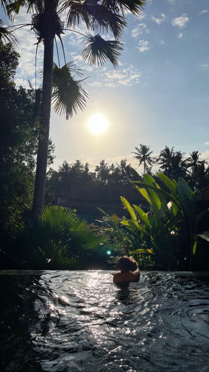 two people are swimming in the pool with palm trees and sun shining through the clouds