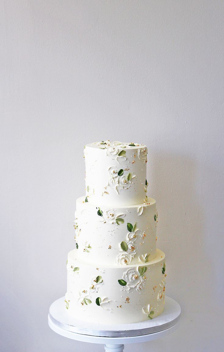 a three tiered cake with white flowers on it is sitting on a stand against a wall