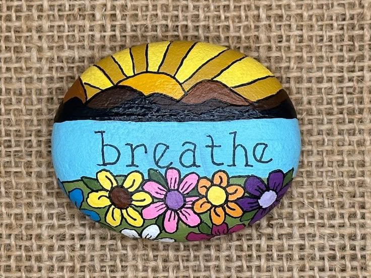 a painted rock that says breathe with flowers and mountains in the background on burlap