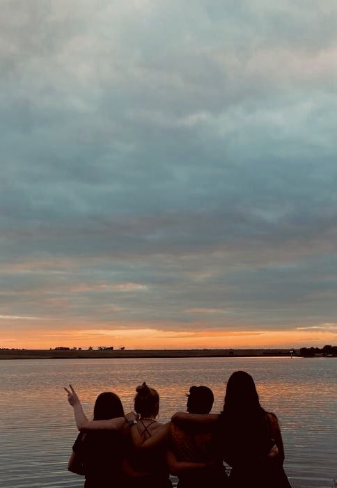 three people sitting on the edge of a body of water with their arms around each other