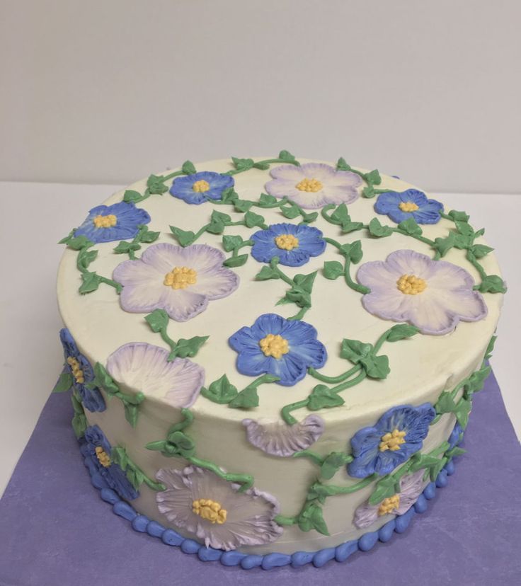 a white cake with blue and purple flowers on it sitting on a purple tablecloth