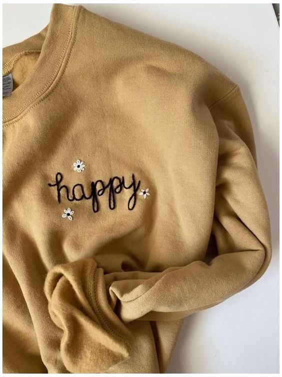 a yellow sweatshirt with the words happy written on it and flowers in black ink, sitting on top of a white surface