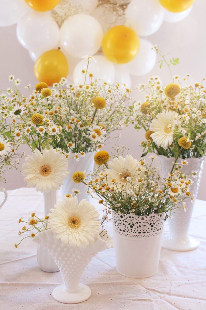 white vases filled with yellow and white flowers on top of a table next to balloons