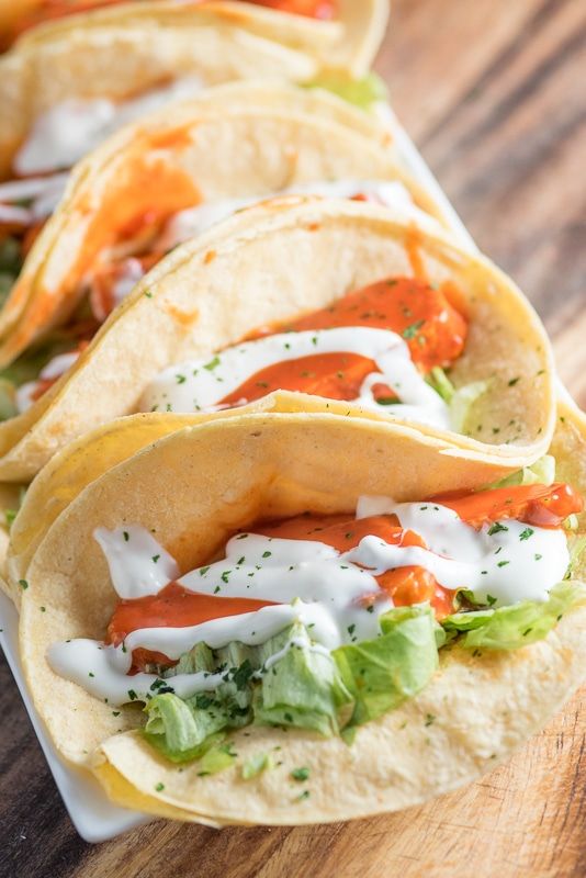 three tacos with sauce and lettuce sitting on a white plate next to a wooden table