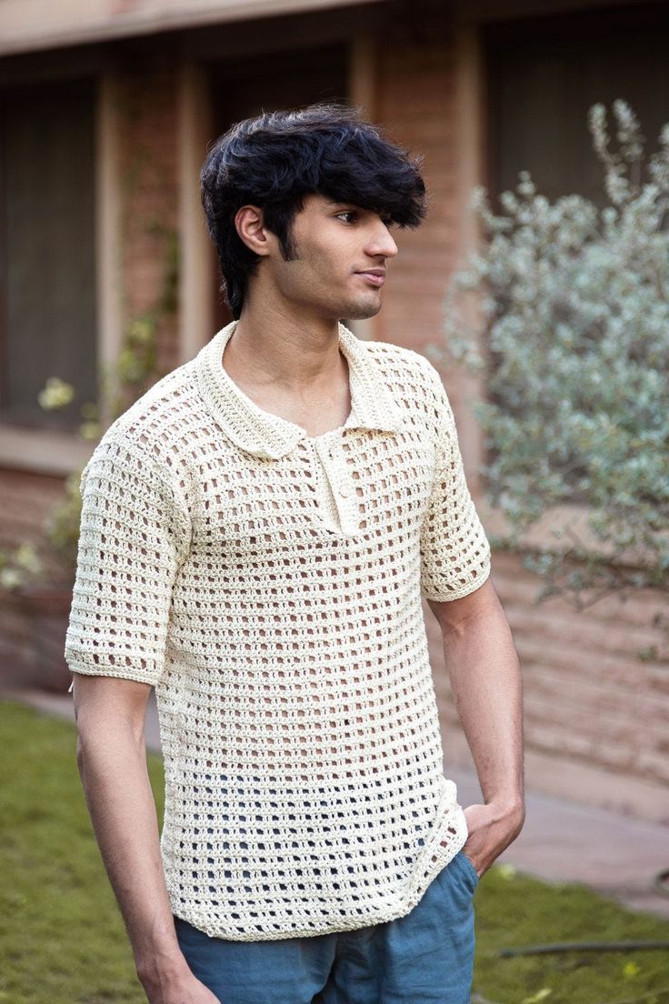 a man standing in front of a house wearing a white crochet shirt and blue jeans