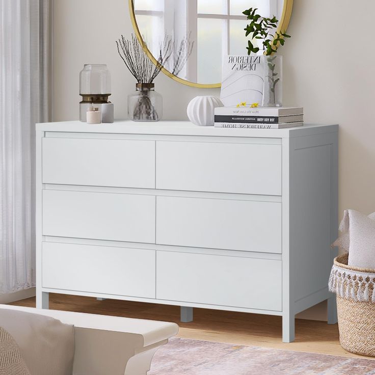 a white dresser sitting next to a window with a round mirror on top of it