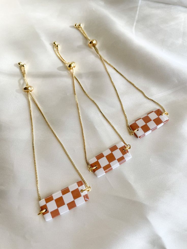 three necklaces with orange and white checkered squares hanging from gold chains on a white sheet