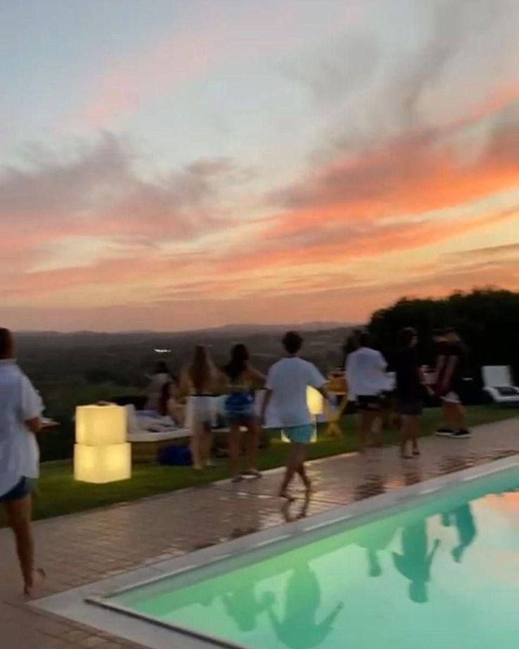 a group of people standing next to a swimming pool at sunset or dawn with the sun setting in the distance