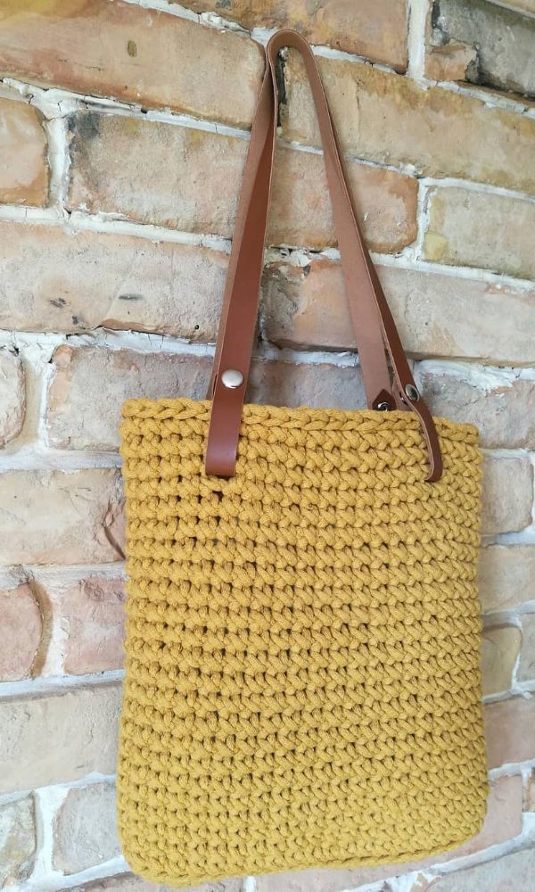 a yellow crochet bag hanging on a brick wall with brown leather handles and straps