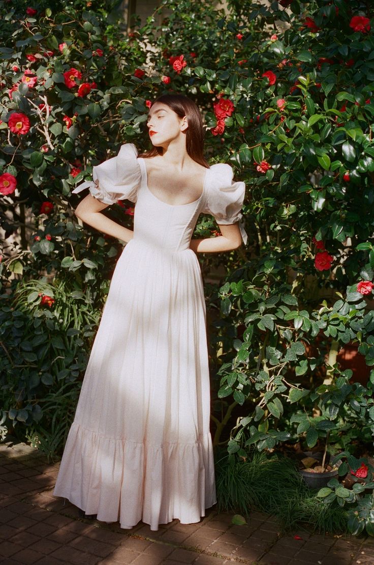 a woman in a white dress standing next to some red flowers and greenery with her hands on her hips