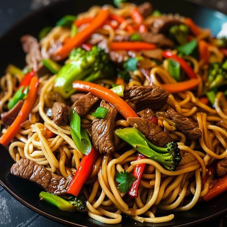 a black plate topped with beef and noodles covered in broccoli, peppers and carrots