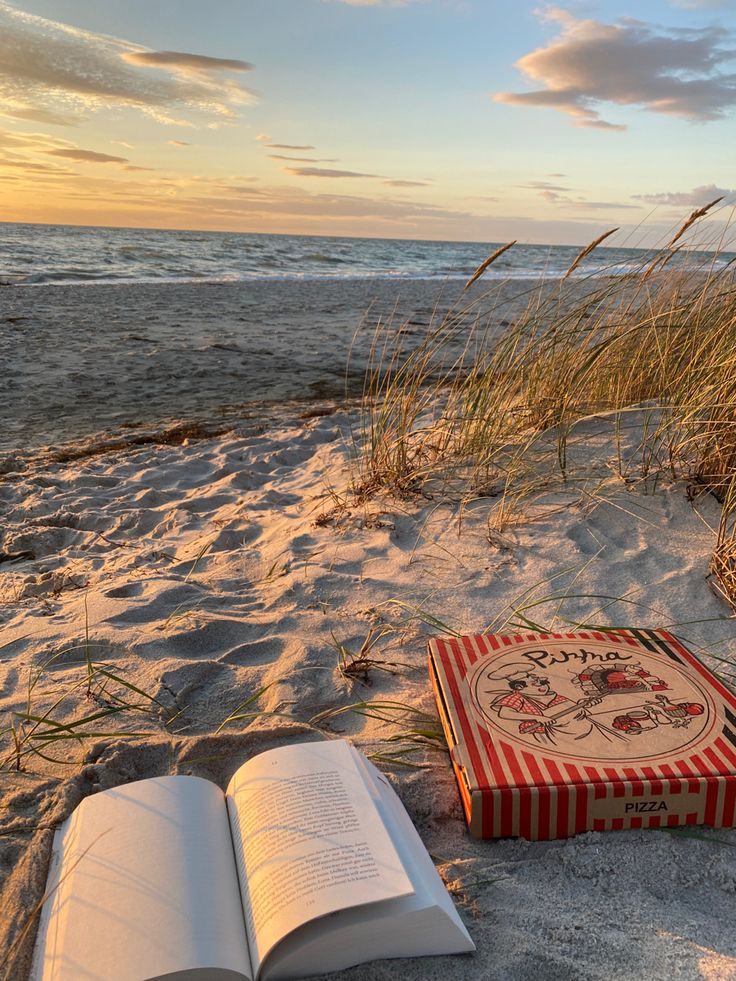 an open book sitting on top of a sandy beach next to the ocean at sunset