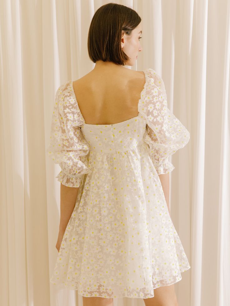 the back of a woman wearing a white dress with yellow flowers on it, standing in front of a curtain