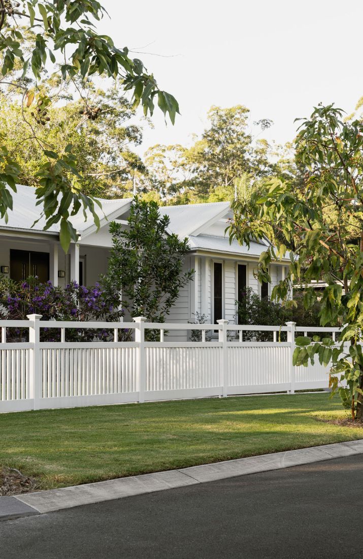 a white picket fence in front of a house with trees and bushes on the side