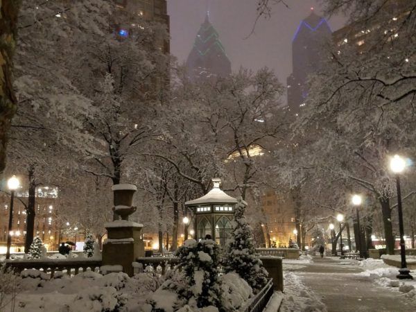 a park covered in snow and surrounded by tall buildings at night with street lamps on either side