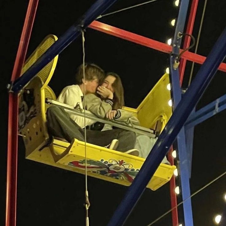 a man and woman sitting on a swing ride at night with lights in the background