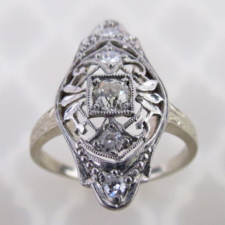 An old European cut diamond is centered on this antique 14k white gold filigree ring. The 3mm .10 carat precious gem is mounted in a square setting. Delicate airy scroll filigree with leaves and millgrain dots decorates the ring. The 13/16 inch long face has a domed gallery with engraved profiles, which is emulated on the ring's shoulders. There are four additional 1.5mm to 1.8mm diamonds set around on the ring. Collectively, this ring has an estimated total weight of .18 cwt diamonds.  The insi Vintage White Gold Jewelry With Diamond Accents, Ornate Diamond-cut Filigree Ring, Vintage White Diamond Filigree Ring, Antique Diamond White Ring With Diamond Accents, Vintage White Gold Diamond Cut Ring, Victorian Silver Filigree Ring With Diamond Accents, Vintage Silver Filigree Ring With Diamond Accents, Vintage White Filigree Ring With Diamond Cut, White Vintage Filigree Ring With Diamond Cut