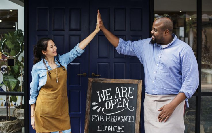 two people standing in front of a restaurant holding their hands up to the sign that says we are open for brunch lunch