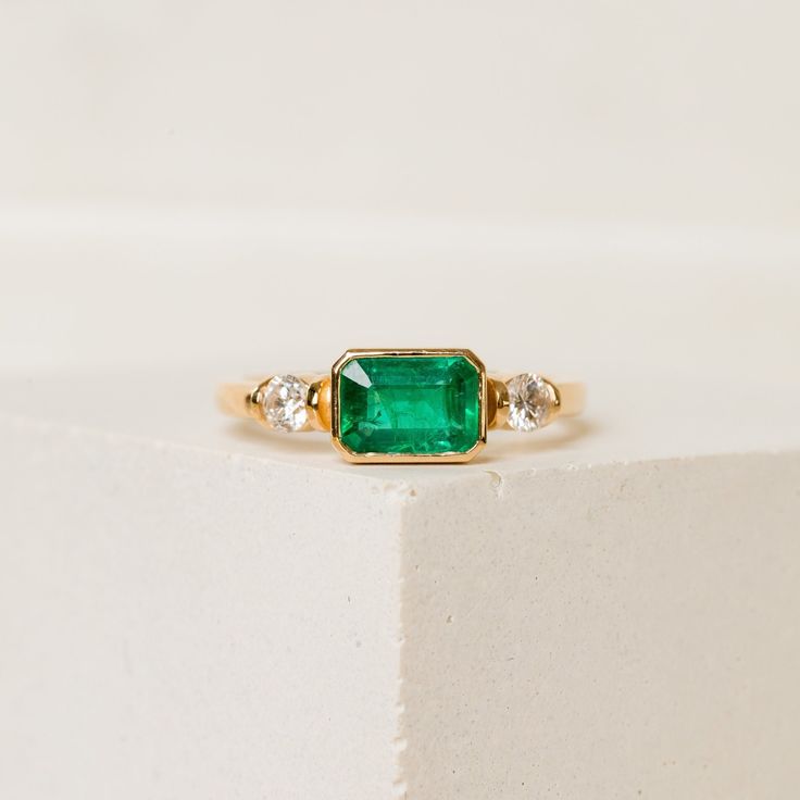 East-West Emerald Engagement Ring An emerald centre stone, set east-west in a 14k yellow gold bezel and flanked by two natural diamonds. The Winona is a three-stone engagement ring with a lot of personality--you'll never tire of her emerald charm.   ORDERING INFORMATION:  From the drop-down menu, please select your choice of centre stone and gold type. In the message to the seller box during the checkout, please let us know the ring size. US 2-12. We offer sizes in 0.25 increments. CENTRE STONE: Emerald Ring With Diamond Band, Bezel 3 Stone Ring, Green Three Stone Birthstone Ring In 14k Gold, Heirloom Green Diamond Ring With Bezel Setting, Heirloom Emerald Ring With Bezel Setting For May Birthstone, Heirloom Emerald Ring With Bezel Setting, Green Emerald Three Stone Ring In 14k Gold, Three Stone Emerald Ring In 14k Gold, Heirloom Green Emerald Ring With Bezel Setting