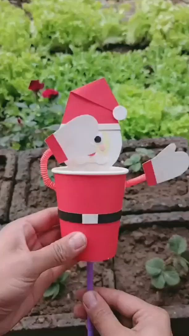 This may contain: someone is holding up a paper cup with santa claus on it in front of some plants