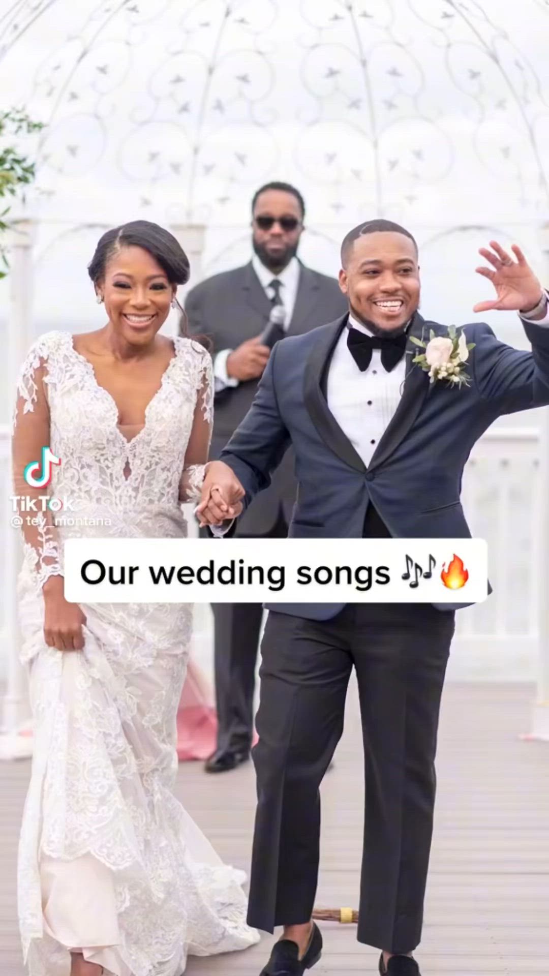 This may contain: a man and woman in formal wear holding hands while walking down a wooden walkway with other people behind them