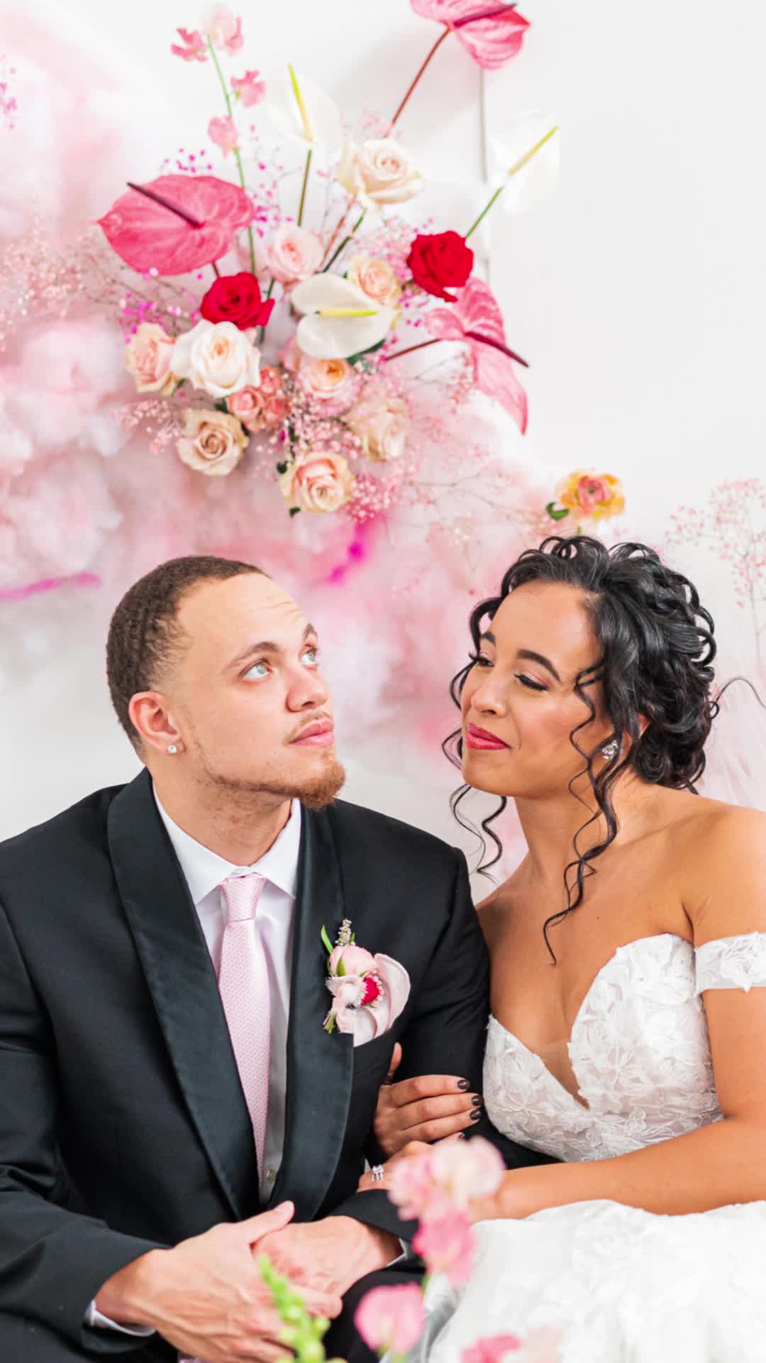 This may contain: a bride and groom sitting next to each other in front of a floral wall decoration