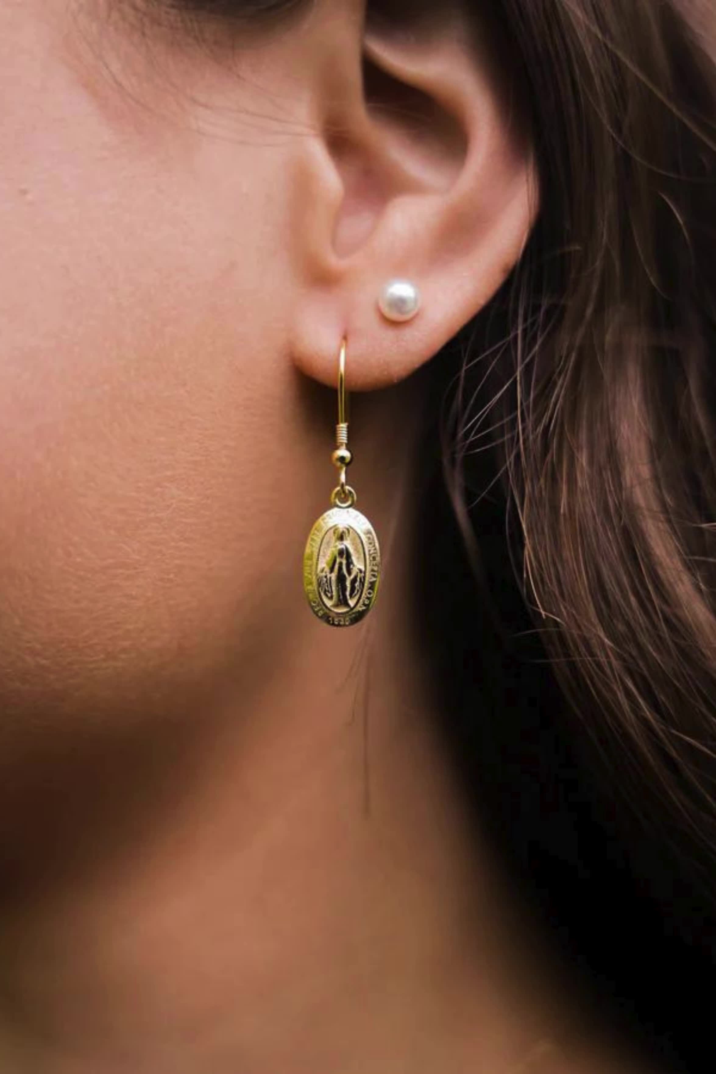 This contains: Close-up of a woman's ear wearing a gold earring with a Miraculous Medal pendant and a small pearl stud.