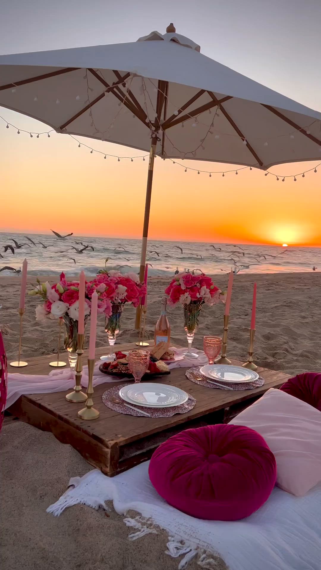 This may contain: a table set up on the beach at sunset with pink flowers and candles in vases