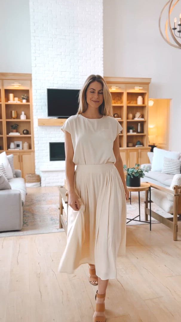 This may contain: a woman standing in a living room wearing a white dress and tan sandals with her hand on her hip