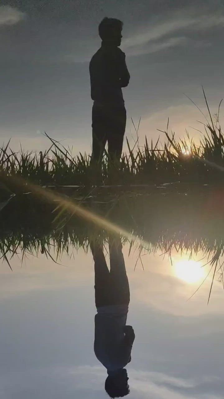 This may contain: a person standing in front of a body of water with the sun setting behind them