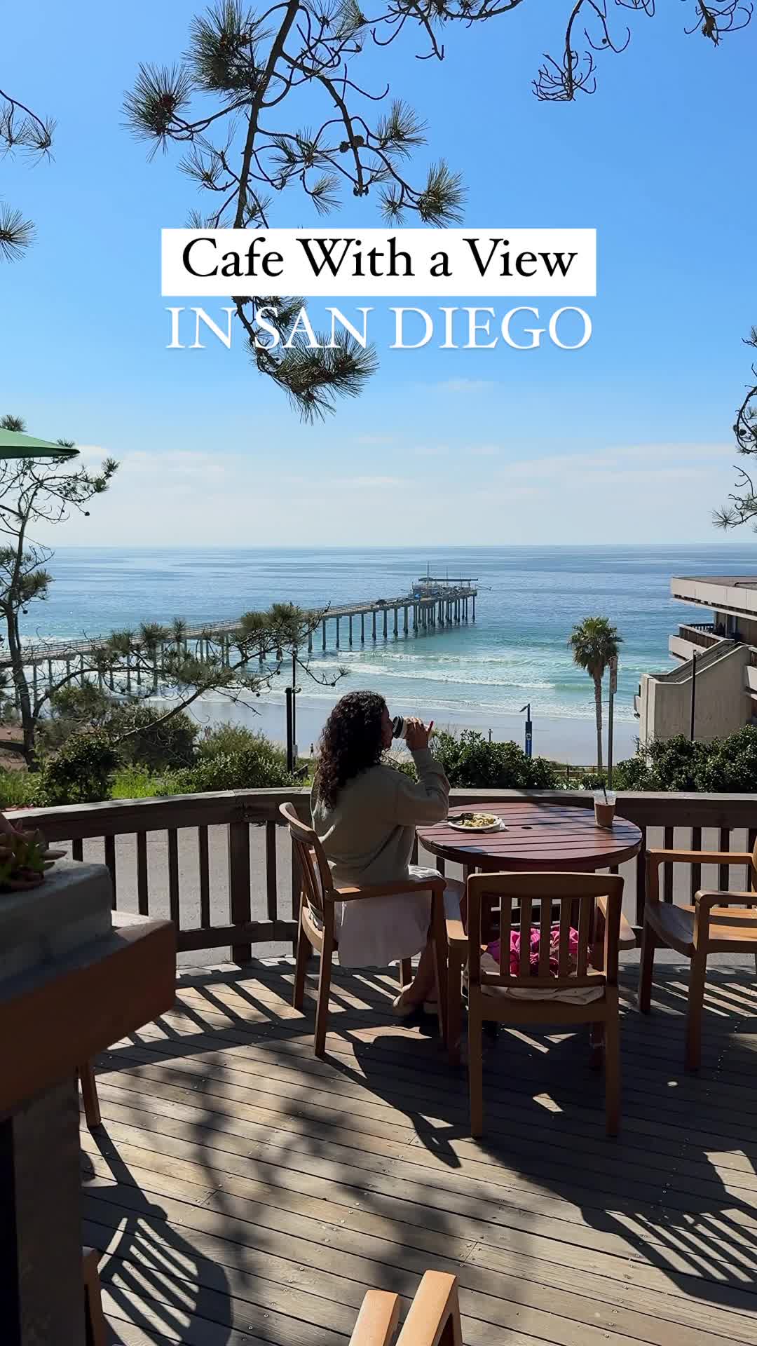 This may contain: a woman sitting at a table on top of a wooden deck next to the ocean