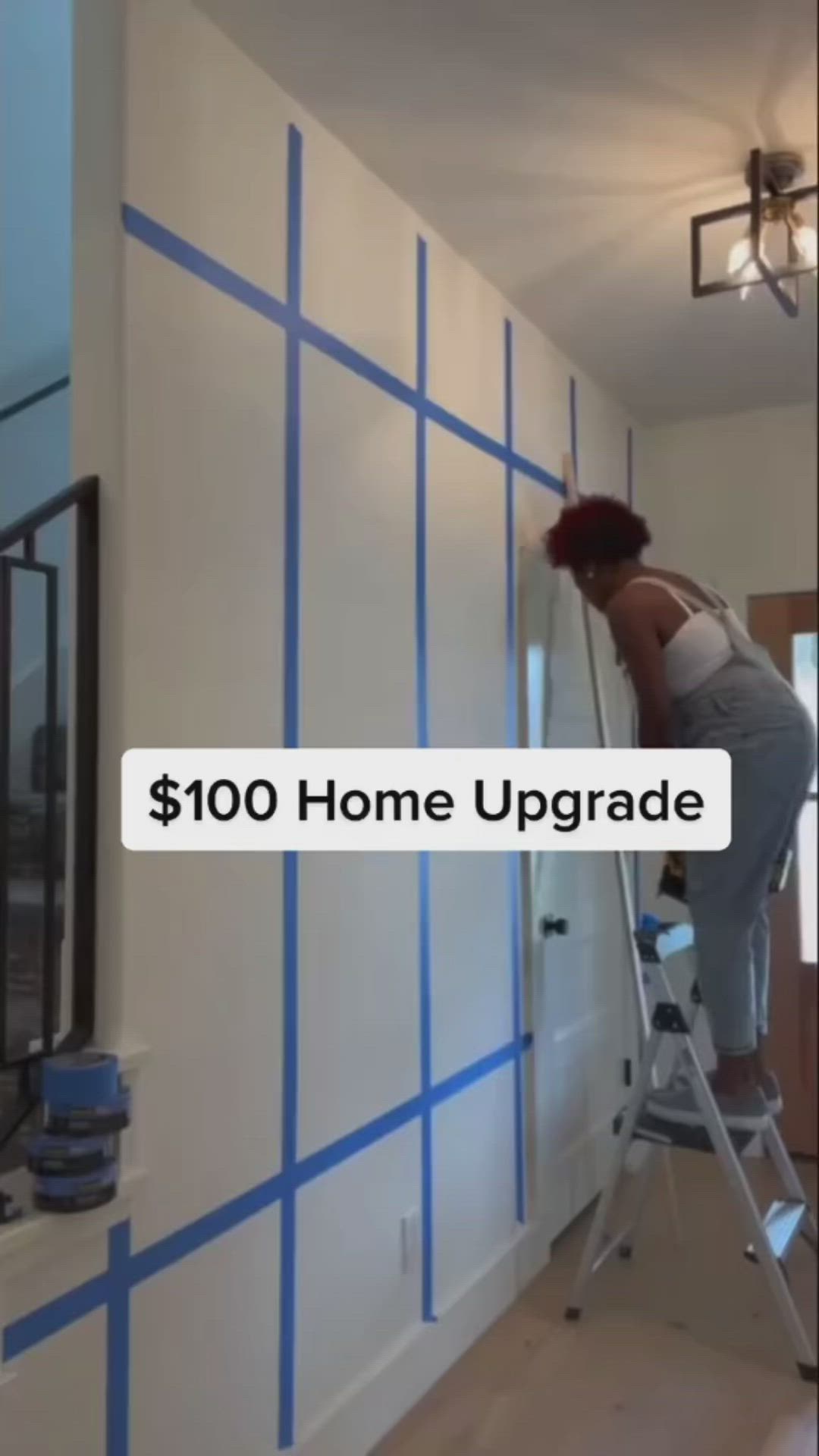 This may contain: a woman on a ladder painting the walls in her living room with blue tape around it