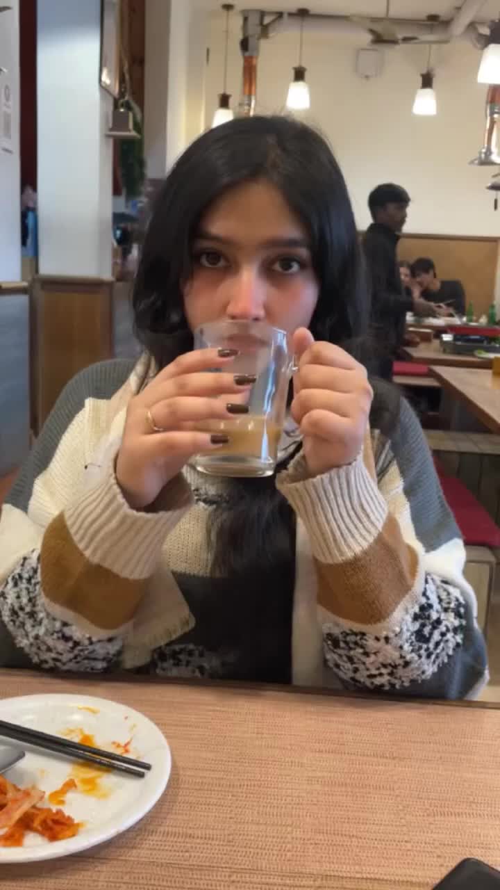 This may contain: a woman sitting at a table drinking from a glass with chopsticks in front of her